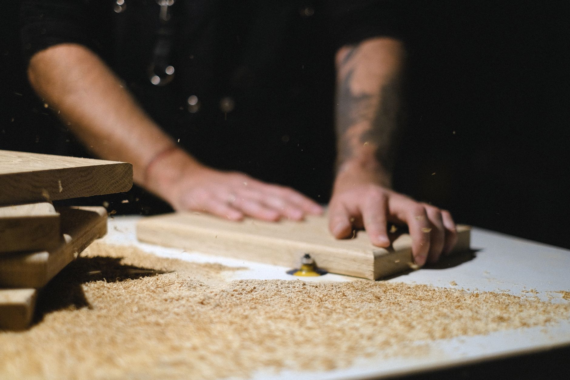 Unrecognizable male master standing at router table while shaping wooden plank at workbench with shavings in dark carpentry on blurred background